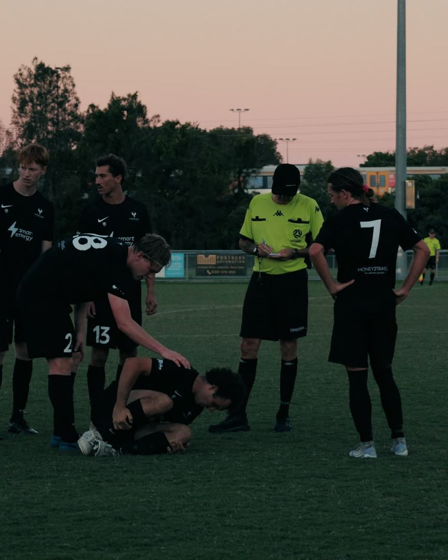 Players and the referee stand over a team-mate on the ground at dusk, game 002.